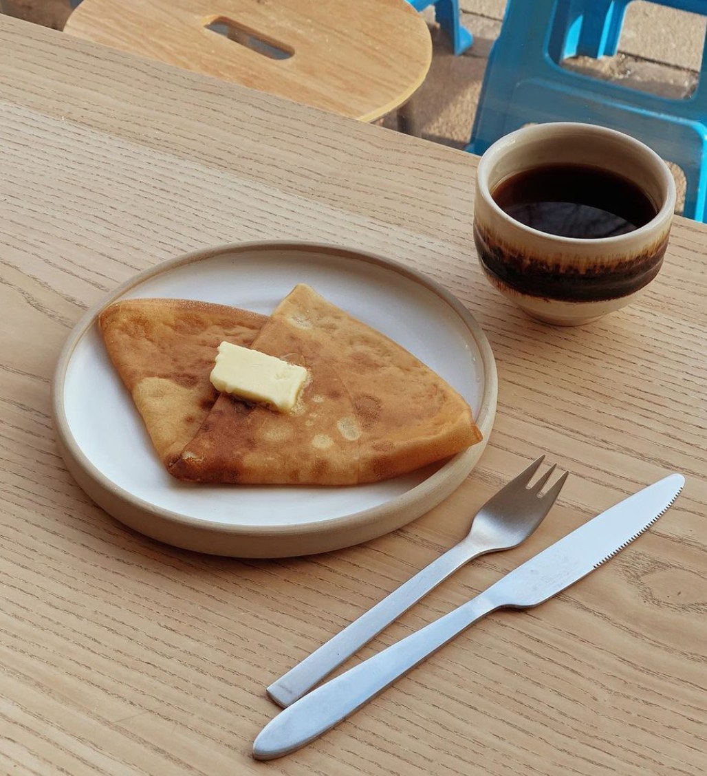木製テーブルに置かれた白い丸皿のバタークレープと茶色い釉薬のコーヒーカップ、青いスツールが背景に見える