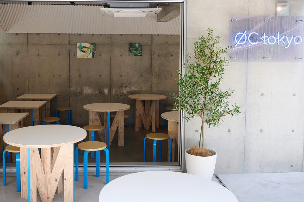 コンクリート壁とブルーのスツール、木製テーブルが並ぶ店内と、右手に光るØC tokyoのネオンサイン、手前にオリーブの鉢植え