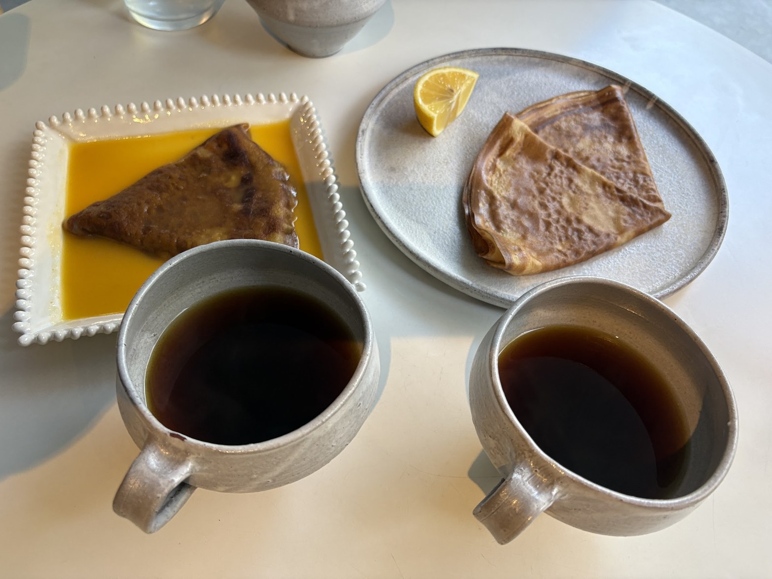 シルバーの陶器マグカップ2杯のコーヒーと、オレンジソースに浸かったクレープ・レモン添えのクレープが白いテーブルに並ぶ