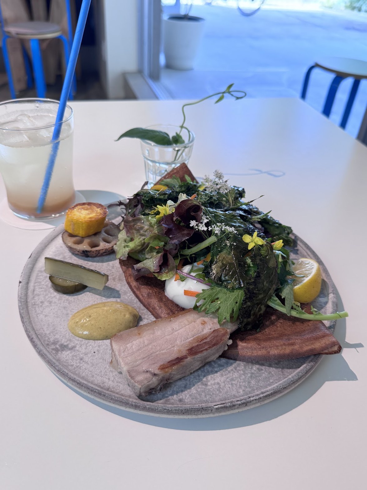 白いテーブルに置かれた大皿に全粒粉クレープ・豚肉・葉野菜・ピクルス・マスタード・レモンが盛られ、左に氷入りのレモネード